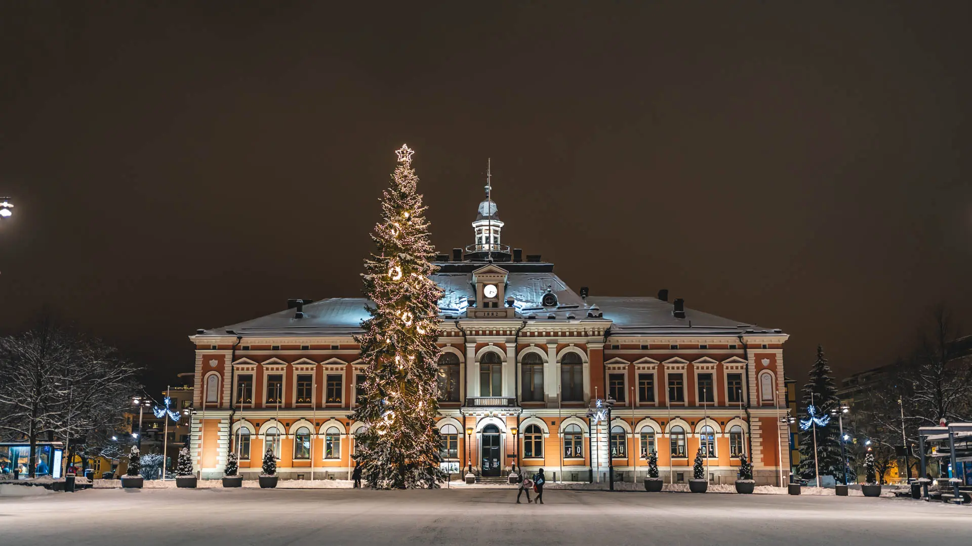 Kuopion kaupungintalo iltavalaistuksessa, edessä joulukuusi koristeltuna valoilla talvisella torilla.