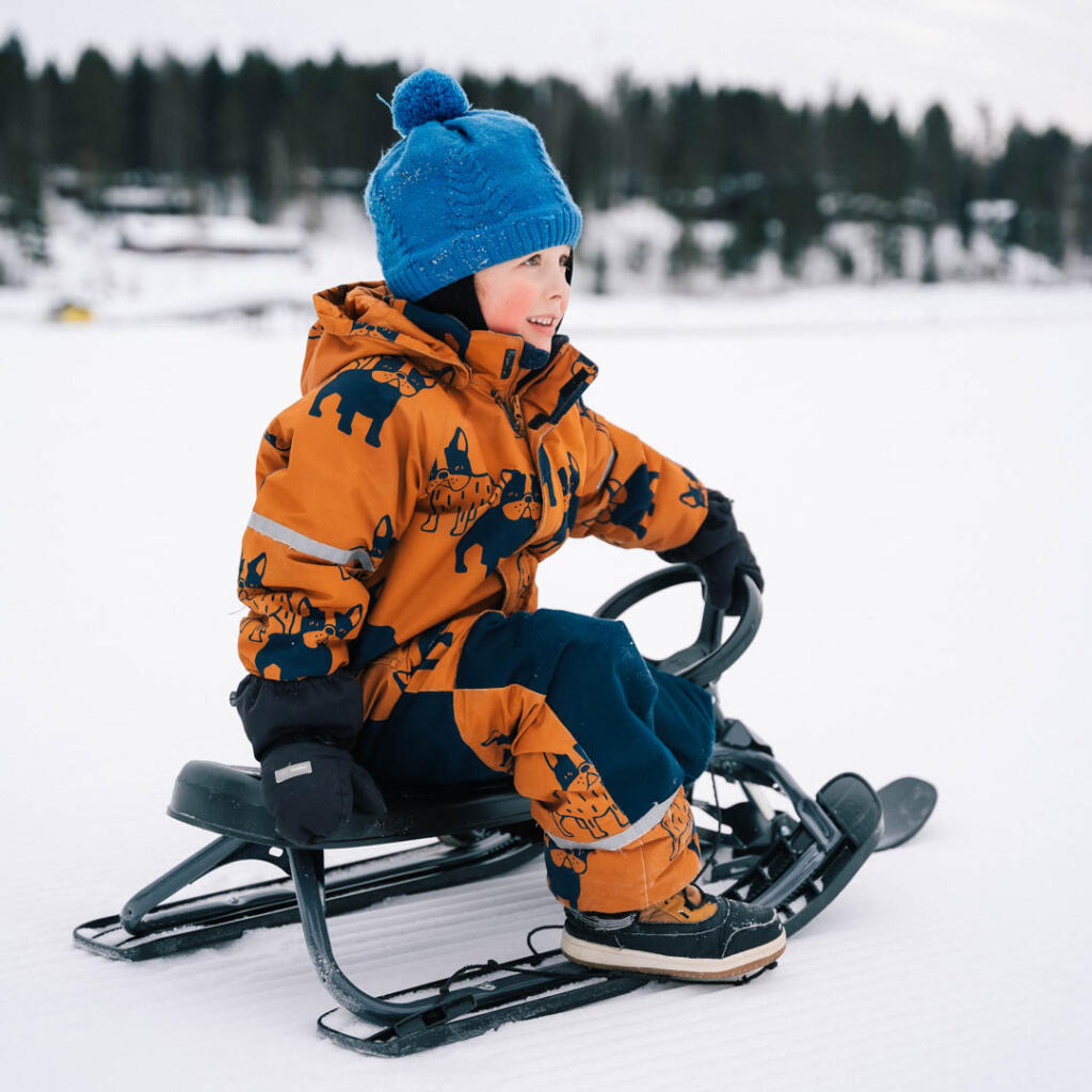 Lapsi istuu potkukelkassa talvisessa maisemassa, pukeutuneena kirkkaan oranssiin haalariin ja siniseen pipoon.